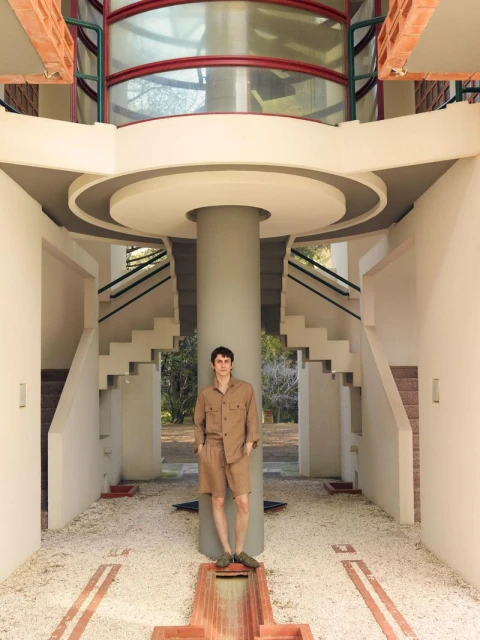 Adult male in casual shirt and shorts poses in the atrium of a modern building with a cylindrical support pillar and stairs.