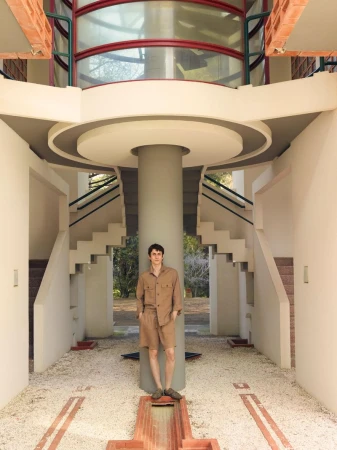 Adult male in casual shirt and shorts poses in the atrium of a modern building with a cylindrical support pillar and stairs.