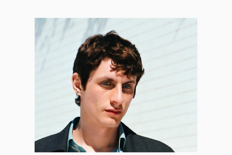 Close-up portrait of a young adult male with wavy brown hair and blue eyes, wearing a blue shirt and black coat, against a white wall.