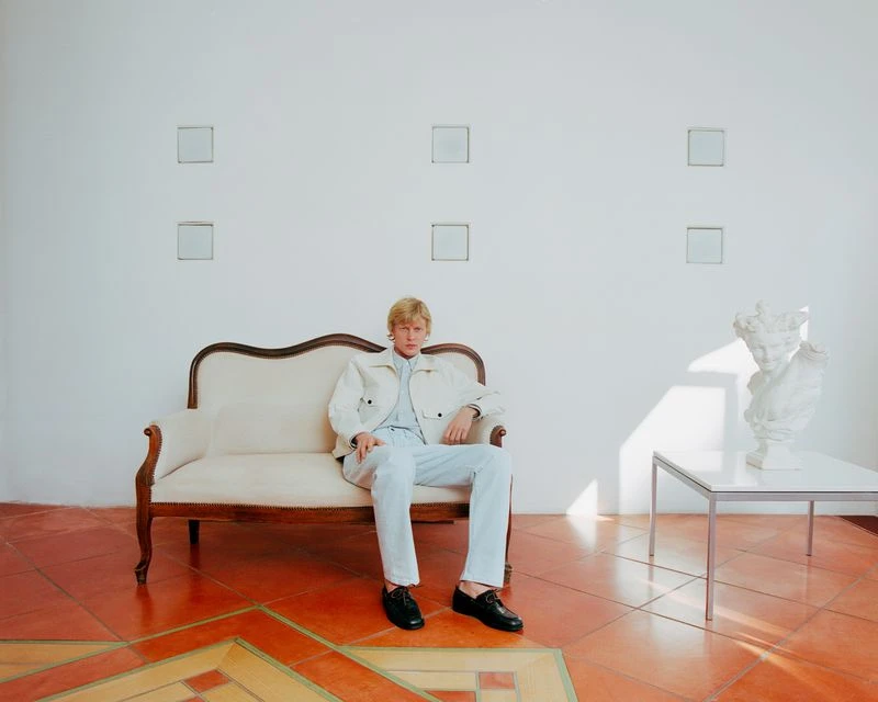 A fair-skinned young adult with blond hair poses sitting on an antique sofa in a white jacket and pants with black shoes, in a studio.