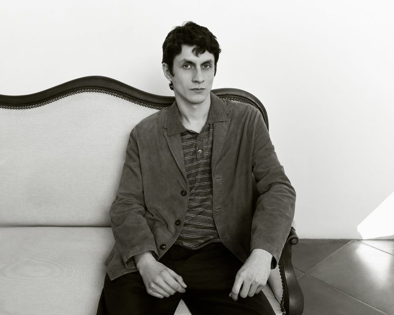 A man with dark hair and a jacket sits on an ornate sofa in a monochrome studio portrait, looking directly at the camera with a neutral expression.
