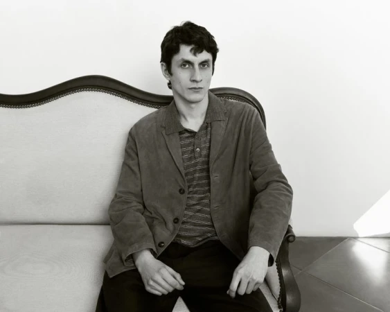 A man with dark hair and a jacket sits on an ornate sofa in a monochrome studio portrait, looking directly at the camera with a neutral expression.