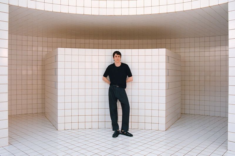 Adult male poses in a black T-shirt and dark pants against a tiled wall and floor in a curved architectural space.