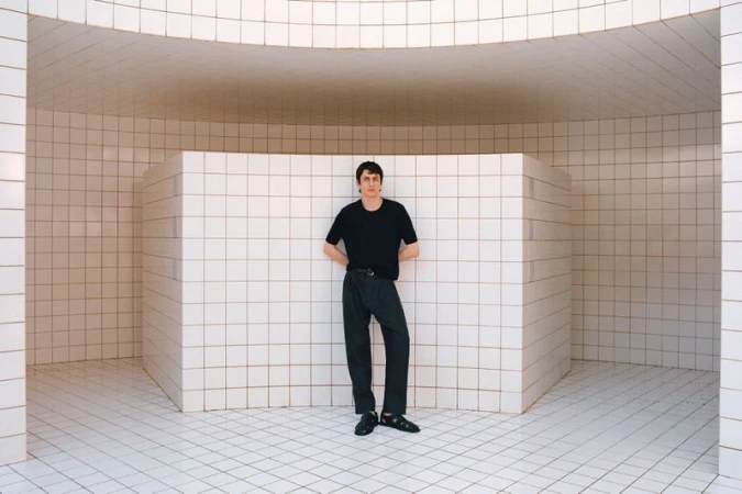 Adult male poses in a black T-shirt and dark pants against a tiled wall and floor in a curved architectural space.