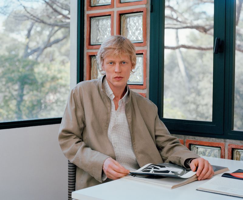 Blonde man wearing a jacket seated at a table with an open book, posed near a window with a view of trees, indoor setting.