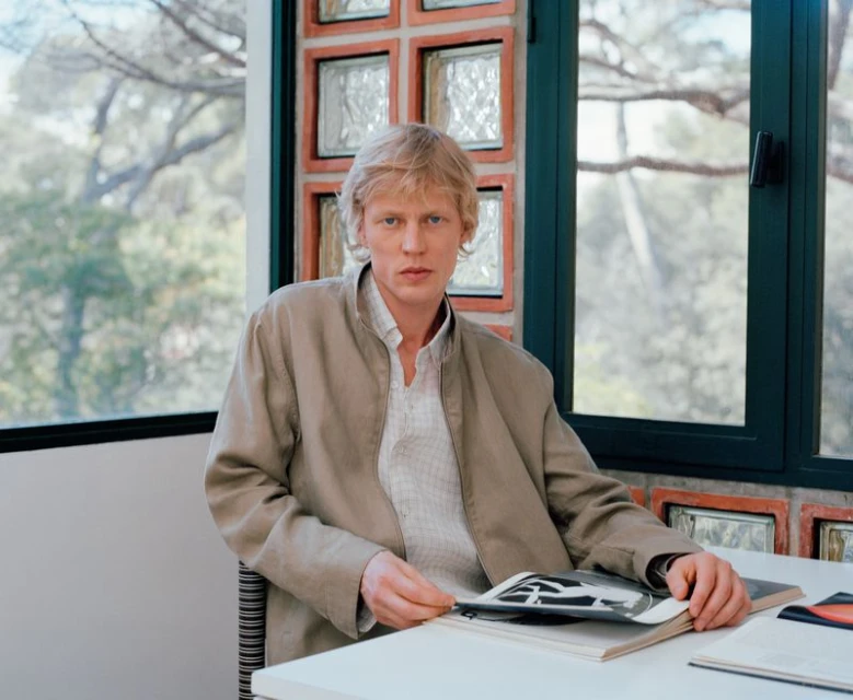 Blonde man wearing a jacket seated at a table with an open book, posed near a window with a view of trees, indoor setting.