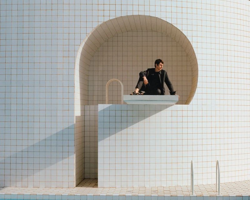 Adult male in a dark suit sitting on a platform in front of an arched window opening in a tiled wall, with a swimming pool nearby.