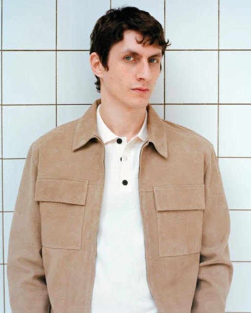 Close-up studio shot of a young adult male wearing a tan suede jacket and cream polo shirt against a white tiled background.