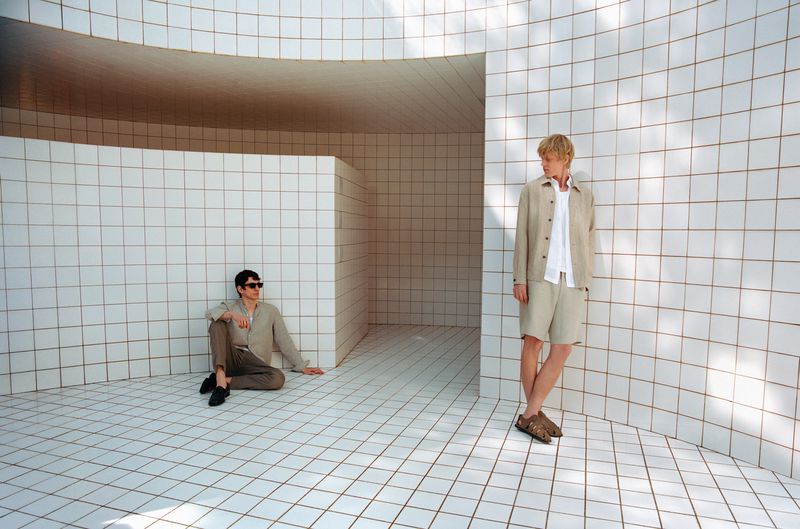 Two men pose in coordinated beige outfits against a geometric white-tiled interior, one seated and the other standing.