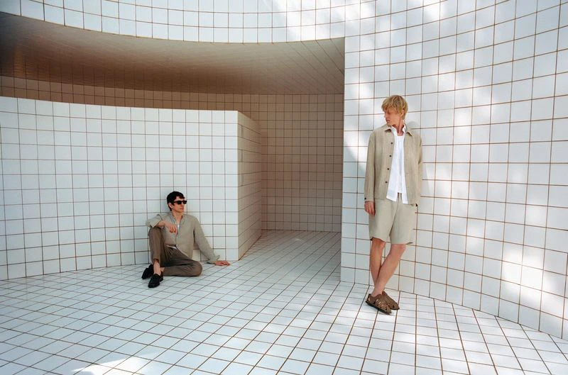 Two men pose in coordinated beige outfits against a geometric white-tiled interior, one seated and the other standing.