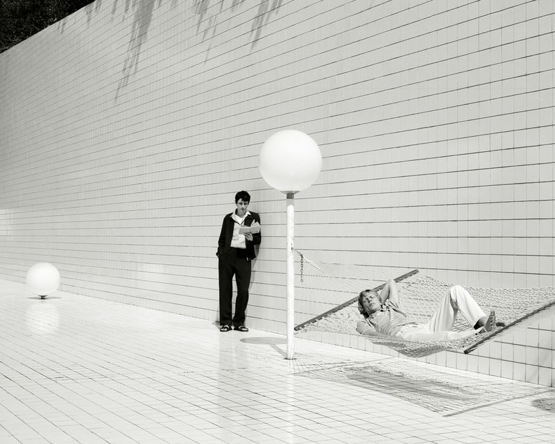 Monochrome outdoor shot of two men: one lying in a hammock, the other leaning against a tiled wall reading a book.