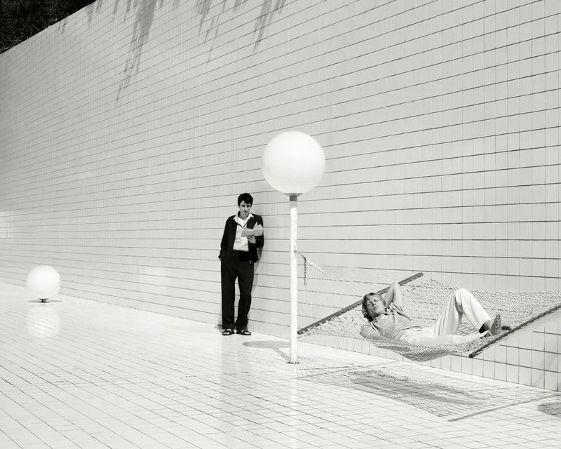 Monochrome outdoor shot of two men: one lying in a hammock, the other leaning against a tiled wall reading a book.