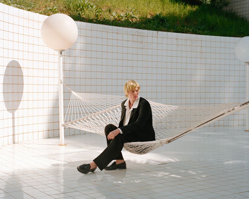A fair-skinned adult sits in a hammock, wearing a black suit and black shoes, surrounded by white tiled walls, and a sphere lamp.