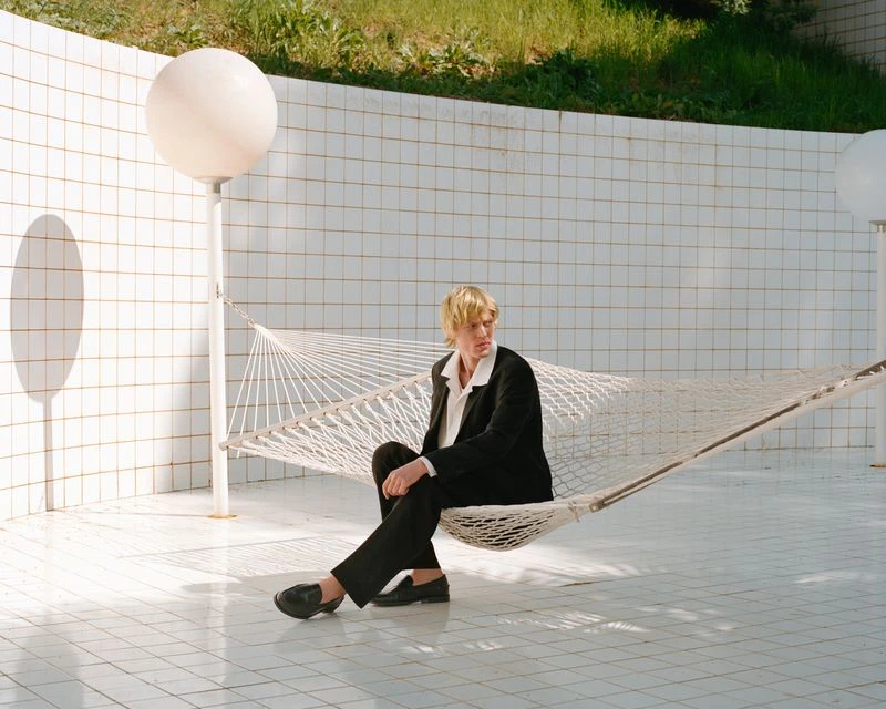 A fair-skinned adult sits in a hammock, wearing a black suit and black shoes, surrounded by white tiled walls, and a sphere lamp.