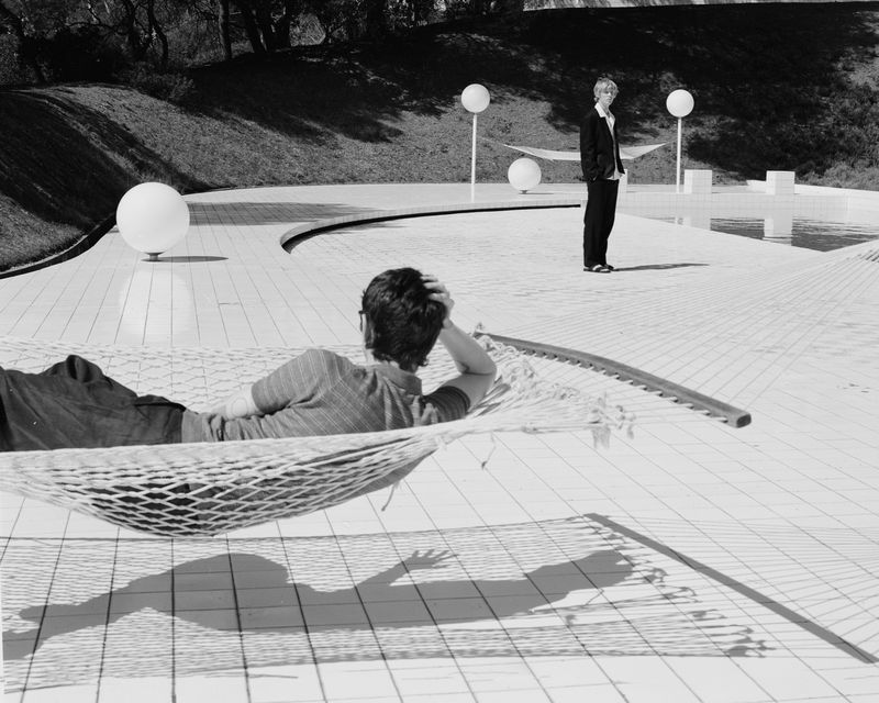 Black and white photo of a man relaxing in a hammock, with another man standing nearby on a white-tiled deck in an outdoor setting.
