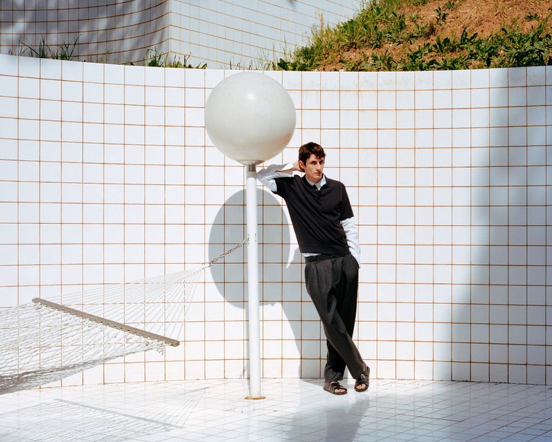 Adult man leans against spherical light on post in minimalist setting, wearing dark polo shirt, trousers, and sandals near hammock.