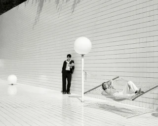 Two models outdoors near a white tiled wall, one standing and reading, the other reclining in a hammock.