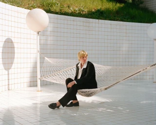 Blonde male model wearing black suit seated in a white net hammock outdoors against tiled walls with spherical lights