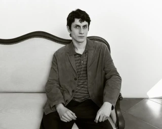 Monochrome portrait of a male model seated on an antique settee wearing a buttoned jacket and striped polo shirt.
