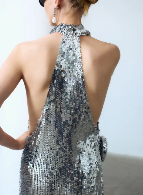 Back view of an adult female with blonde hair posing in a silver sequin halter dress against a white wall, wearing a black eye mask.