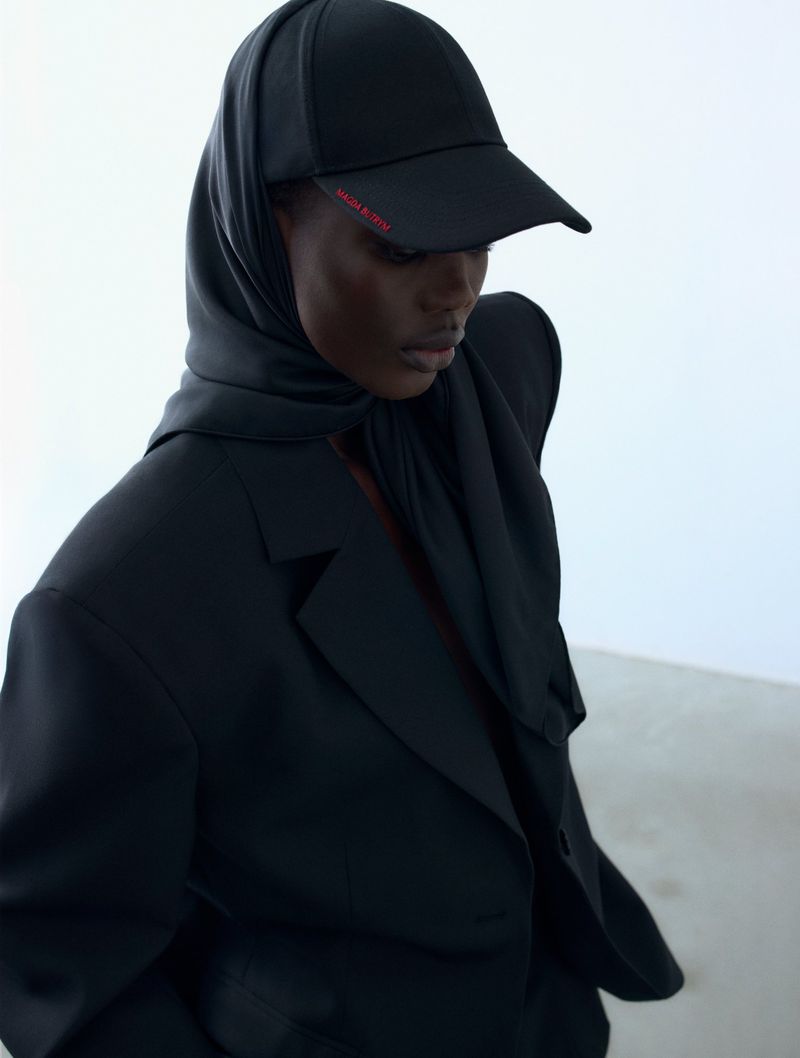 Fashionable woman in a black blazer and cap with headscarf, posing in a minimalist photo studio with bright, even lighting.