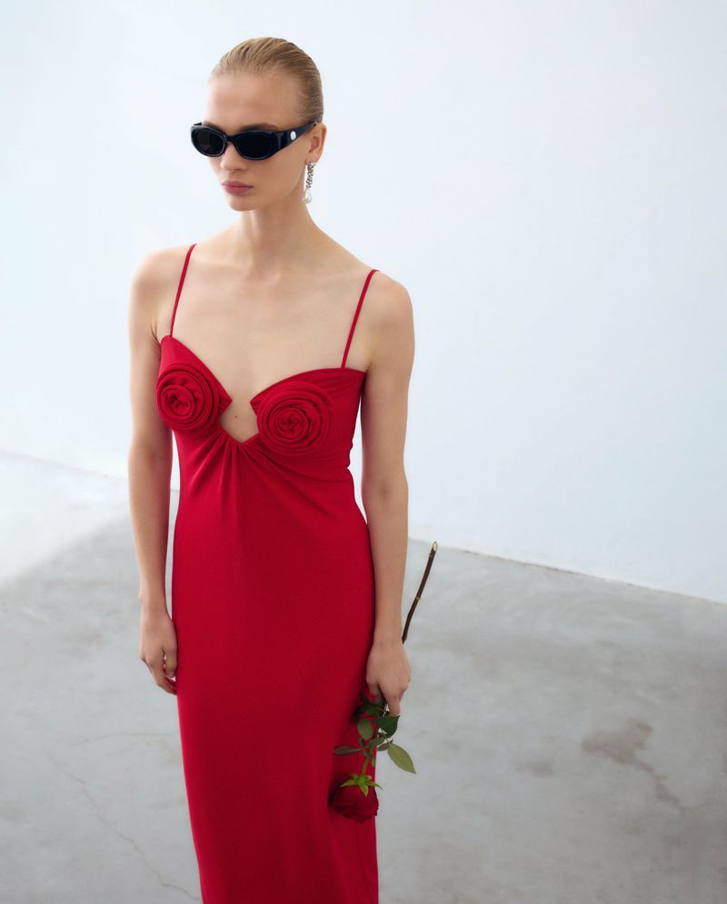A slender woman in a red dress poses in a white studio, holding a rose. She wears black sunglasses and dangling earrings.