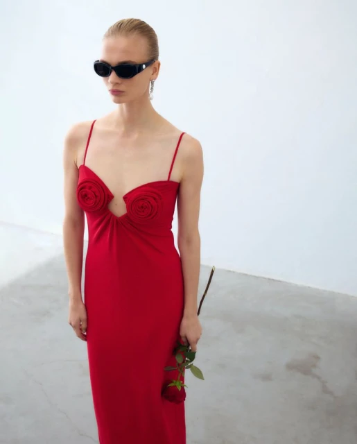A slender woman in a red dress poses in a white studio, holding a rose. She wears black sunglasses and dangling earrings.