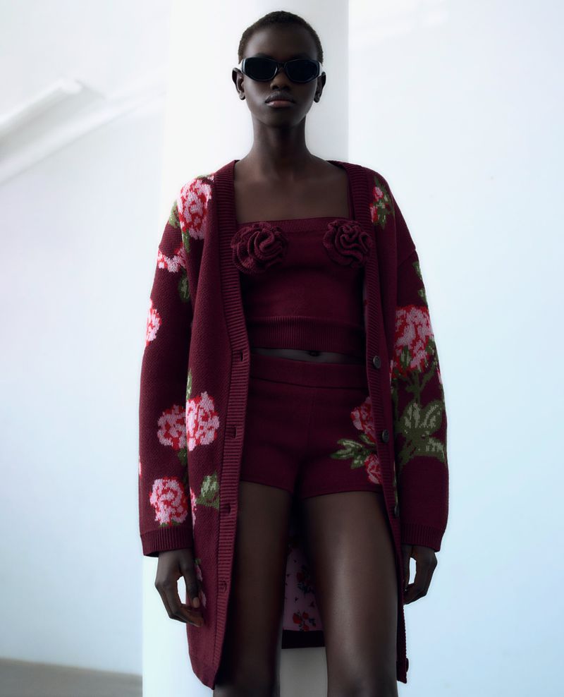 Dark-skinned woman wearing a burgundy floral knitwear ensemble, including a cardigan, top, and shorts, posing indoors against a white wall.