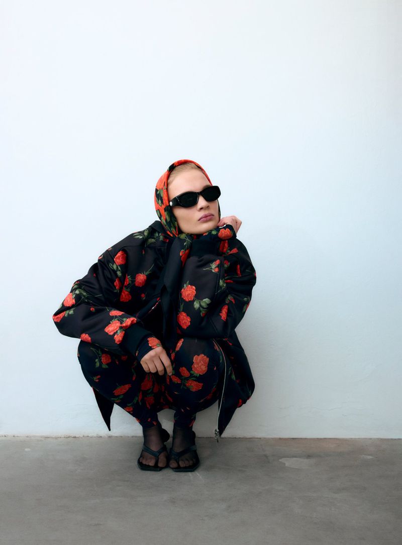 Fashionable adult woman in a rose-patterned jacket, headscarf, leggings, and chunky sunglasses, crouching in front of a white wall.