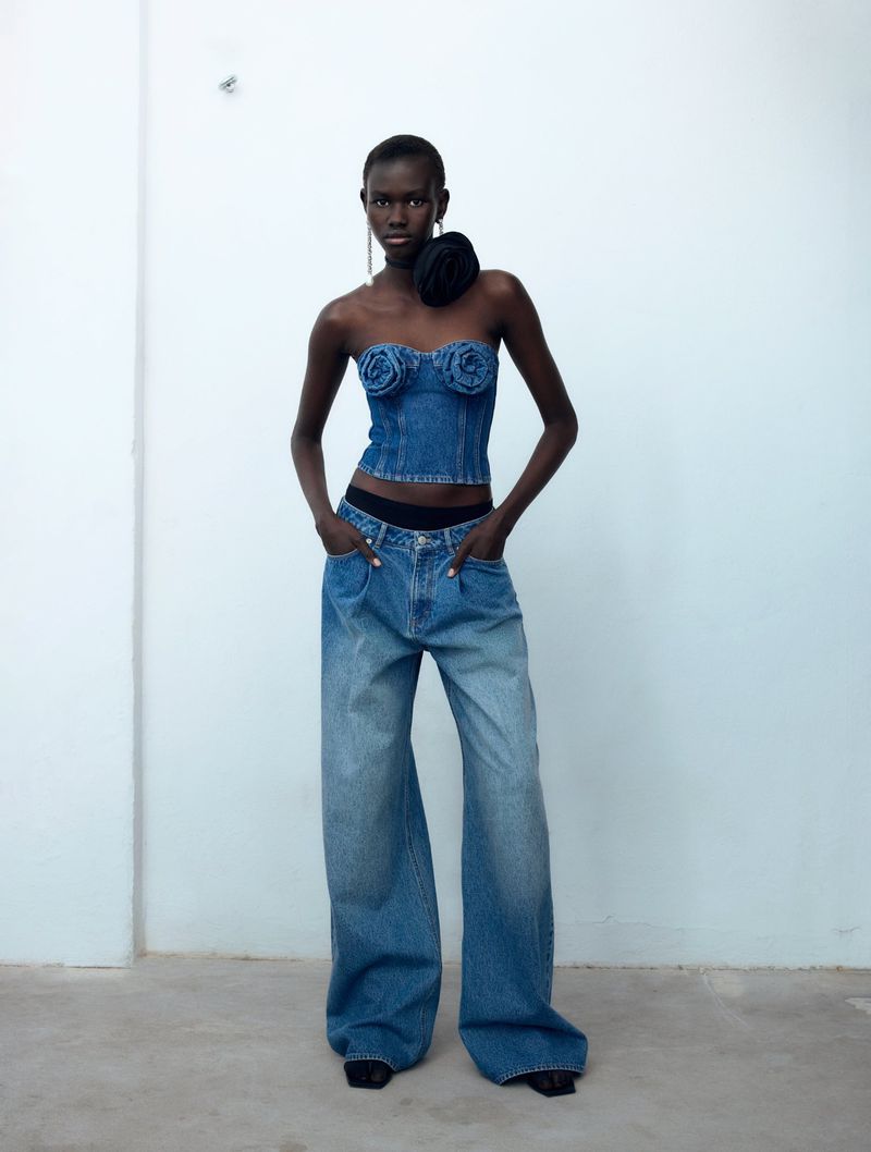 Adult female model wears a denim bustier and baggy jeans in a studio setting with a white wall in the background, fashion studio shot.