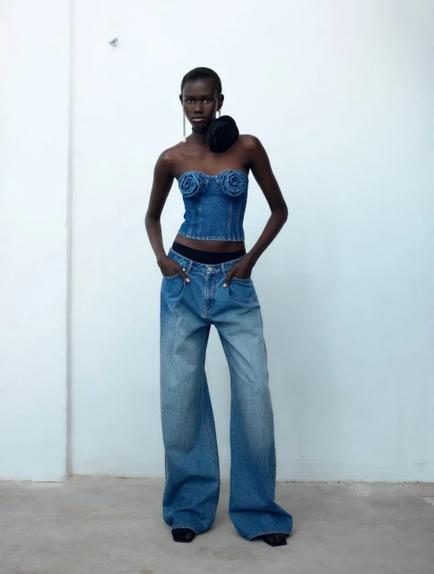 Adult female model wears a denim bustier and baggy jeans in a studio setting with a white wall in the background, fashion studio shot.