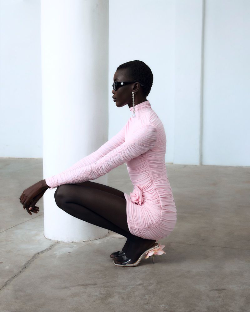 Woman in pink dress poses crouching indoors by white column wearing black sunglasses and black stockings with high heels, fashion shot.