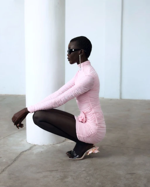 Woman in pink dress poses crouching indoors by white column wearing black sunglasses and black stockings with high heels, fashion shot.