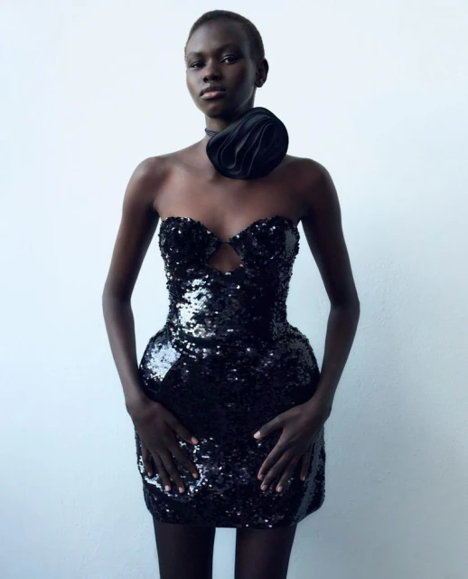 Woman in a black sequin dress with a large flower detail poses against a white wall, showcasing fashion and style.