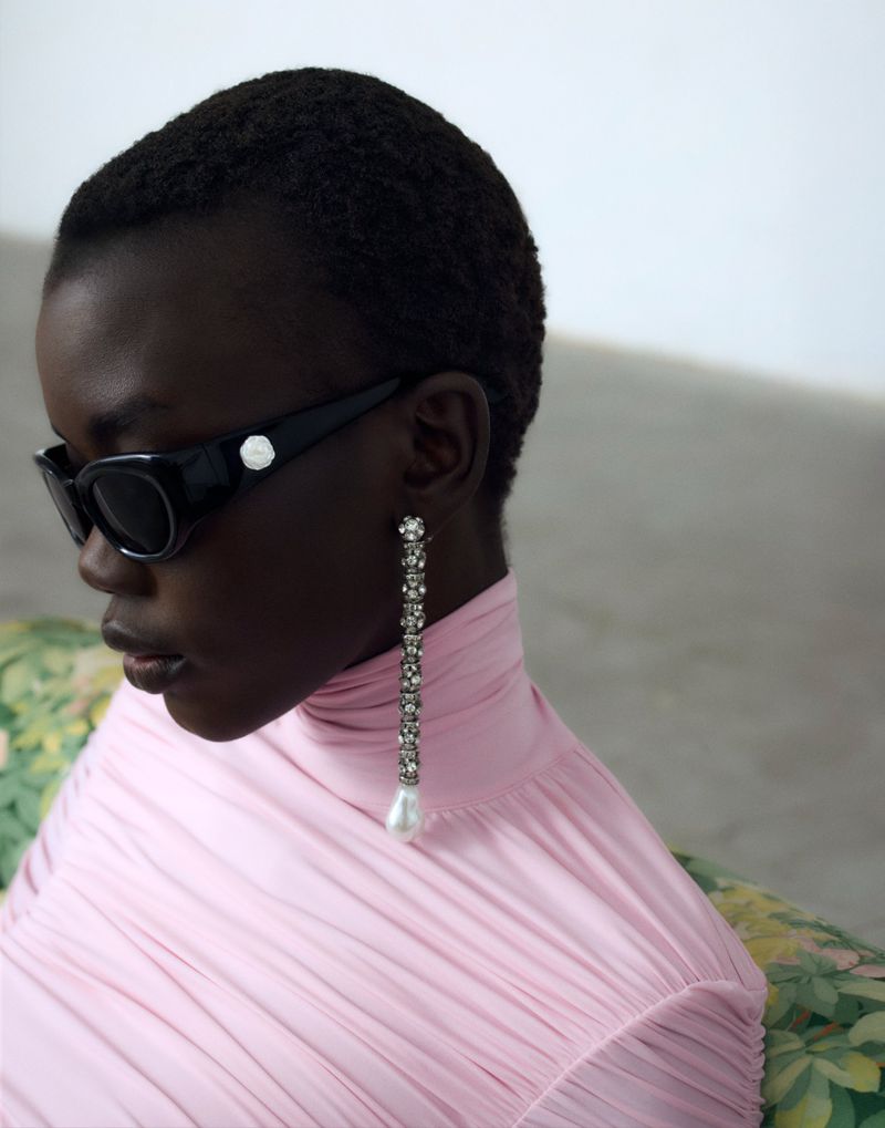 Dark-skinned model wearing sunglasses and a pink dress with a long crystal earring. Studio shot, floral-patterned armchair visible.