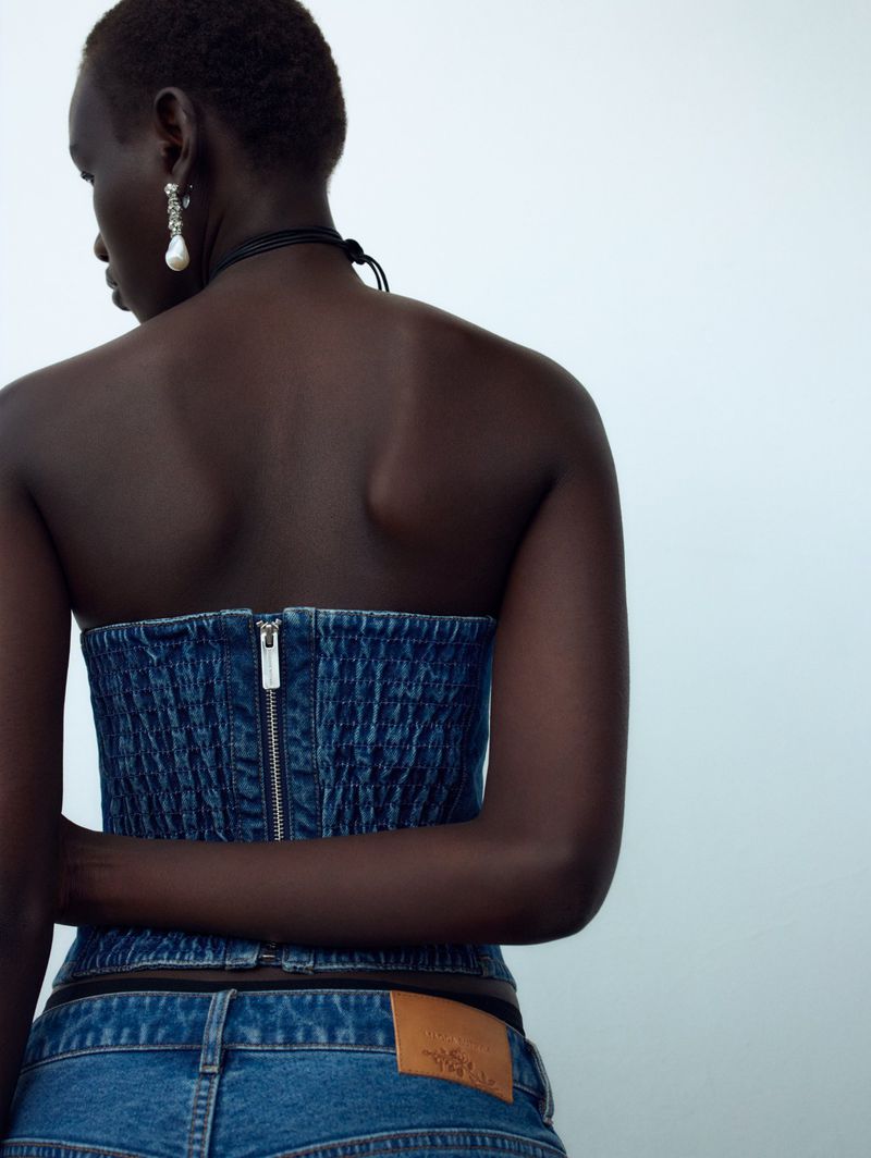 Brunette adult woman poses in a denim corset top and matching jeans against a white background in a fashion photography studio.