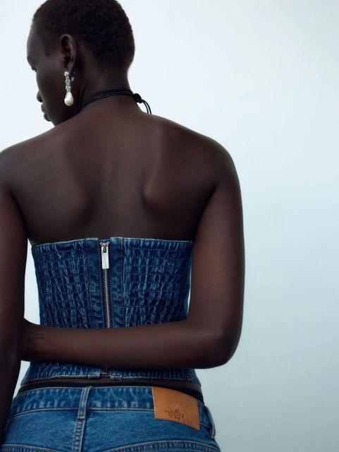 Brunette adult woman poses in a denim corset top and matching jeans against a white background in a fashion photography studio.