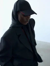 Model in black blazer wearing a black baseball cap over a matching headscarf against a white background.