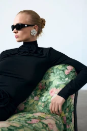 Model wearing black turtleneck and sunglasses seated on floral sofa for H&M x Magda Butrym