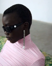 Model wearing dark sunglasses, crystal drop earrings and pink turtleneck against neutral background