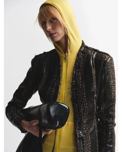 Fashion shot of a blond adult woman wearing a yellow hoodie and black tweed jacket while holding a black patent leather bag.