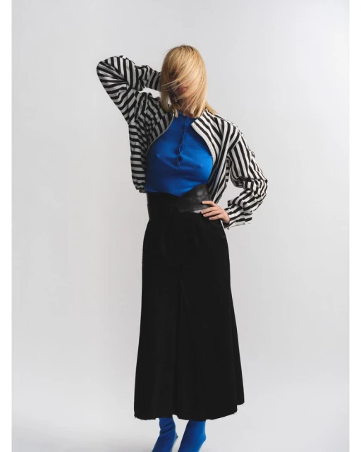Adult woman in a studio wearing black culottes, a blue top, a black and white striped jacket, and blue sock boots poses.