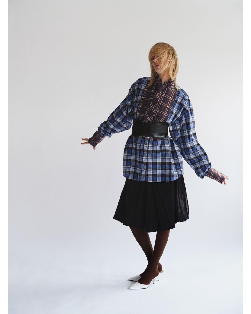 Full-length shot of a woman with blonde hair wearing an oversized blue plaid shirt, black skirt, brown stockings and white heels against a white backdrop.