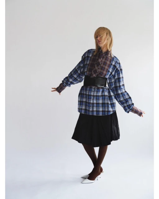 Full-length shot of a woman with blonde hair wearing an oversized blue plaid shirt, black skirt, brown stockings and white heels against a white backdrop.