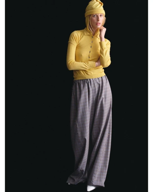 A fashionably dressed young adult poses in a yellow knit top and matching beanie, paired with oversized plaid pants, against a dark background.