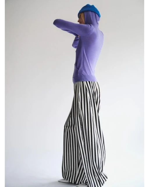 Woman in a light purple hooded sweatshirt and striped palazzo pants stands in a brightly lit white photo studio.