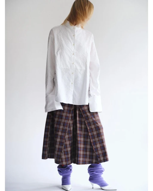 Fashion editorial shot of a woman wearing a white striped shirt, plaid skirt, and purple leg warmers against a white backdrop.