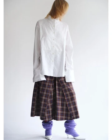Fashion editorial shot of a woman wearing a white striped shirt, plaid skirt, and purple leg warmers against a white backdrop.