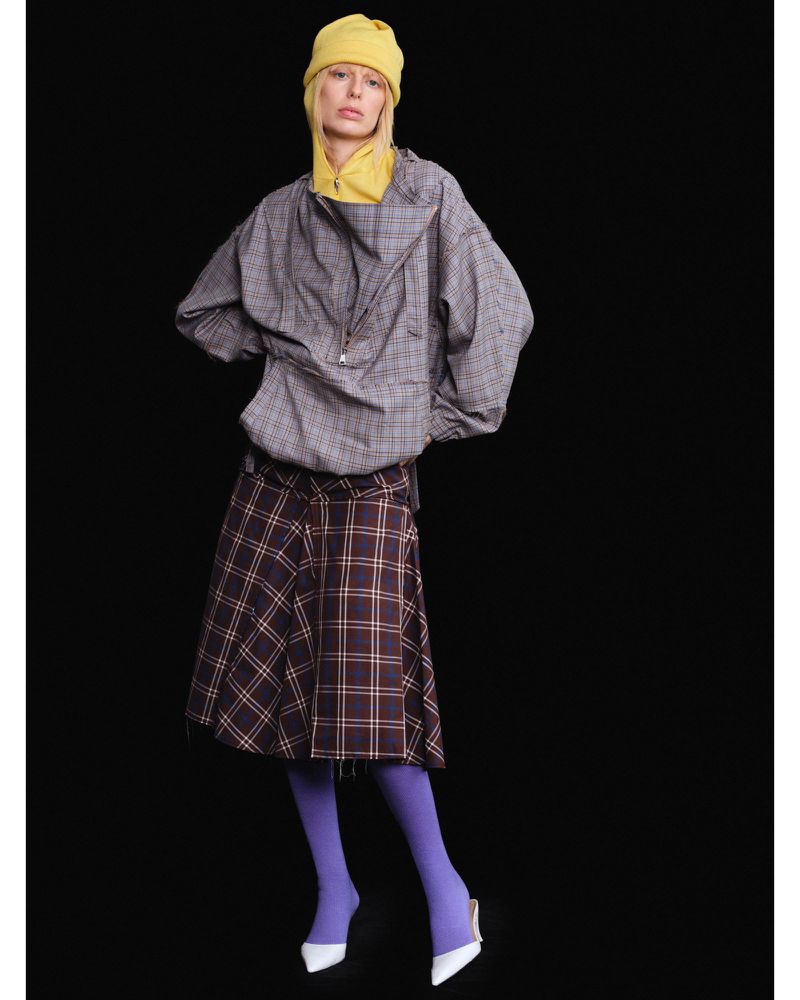 A woman in a yellow knit hat, grey plaid jacket, and plaid skirt poses against a black backdrop for a fashion editorial shot.