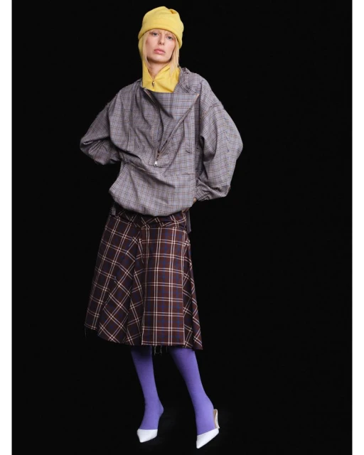 A woman in a yellow knit hat, grey plaid jacket, and plaid skirt poses against a black backdrop for a fashion editorial shot.